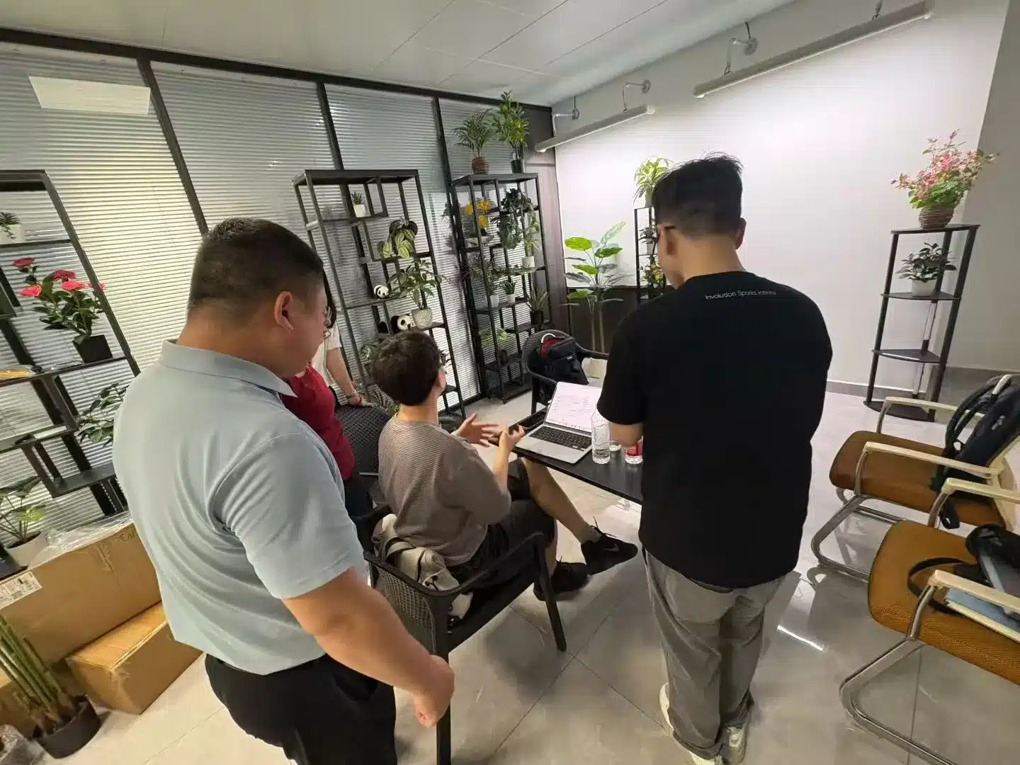 Three Infility specialists gather around a laptop in a modern plant-lined office, reviewing wireframes and analytics for a client website. This hands-on scene illustrates our 外贸独立站定制开发服务 — custom WordPress independent-site development focused on SEO, UX, and lead-generation for B2B exporters.