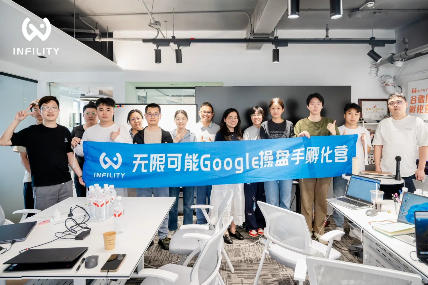 多位学员在明亮培训教室内合影，共同手持印有“无限可能Google操盘手孵化营”字样的蓝色横幅，前方桌面摆放着笔记本电脑与饮用水。