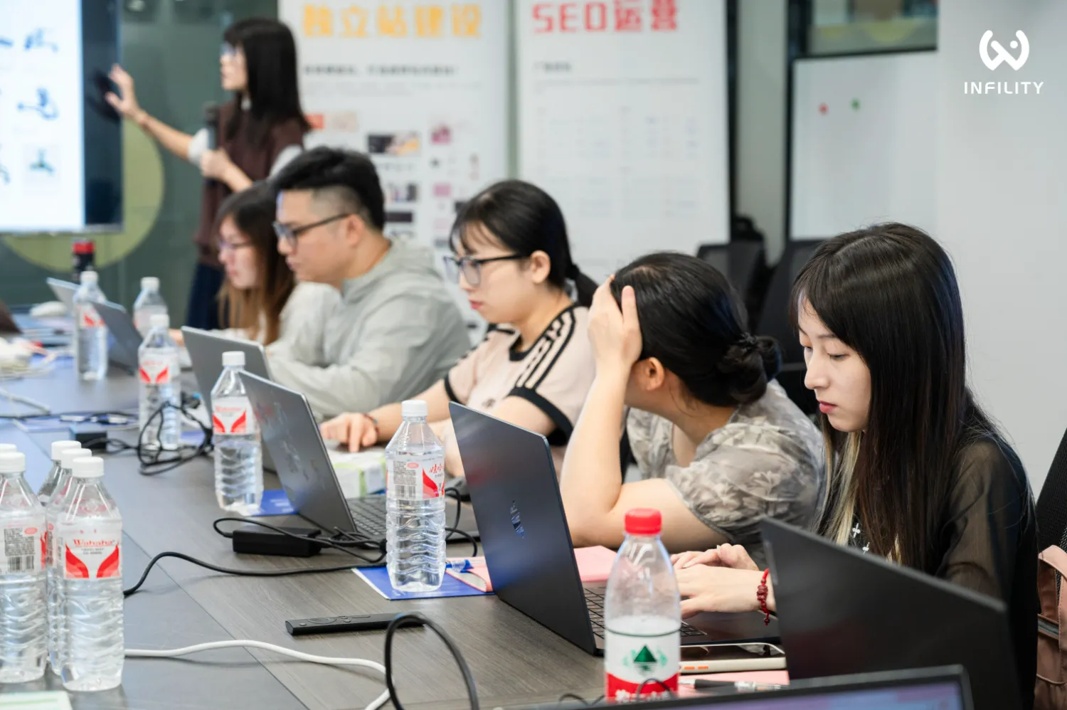 INFILITY举办的独立站建设与SEO运营培训课程现场，学员们正专注地在笔记本电脑上操作学习，讲师在前方进行指导。