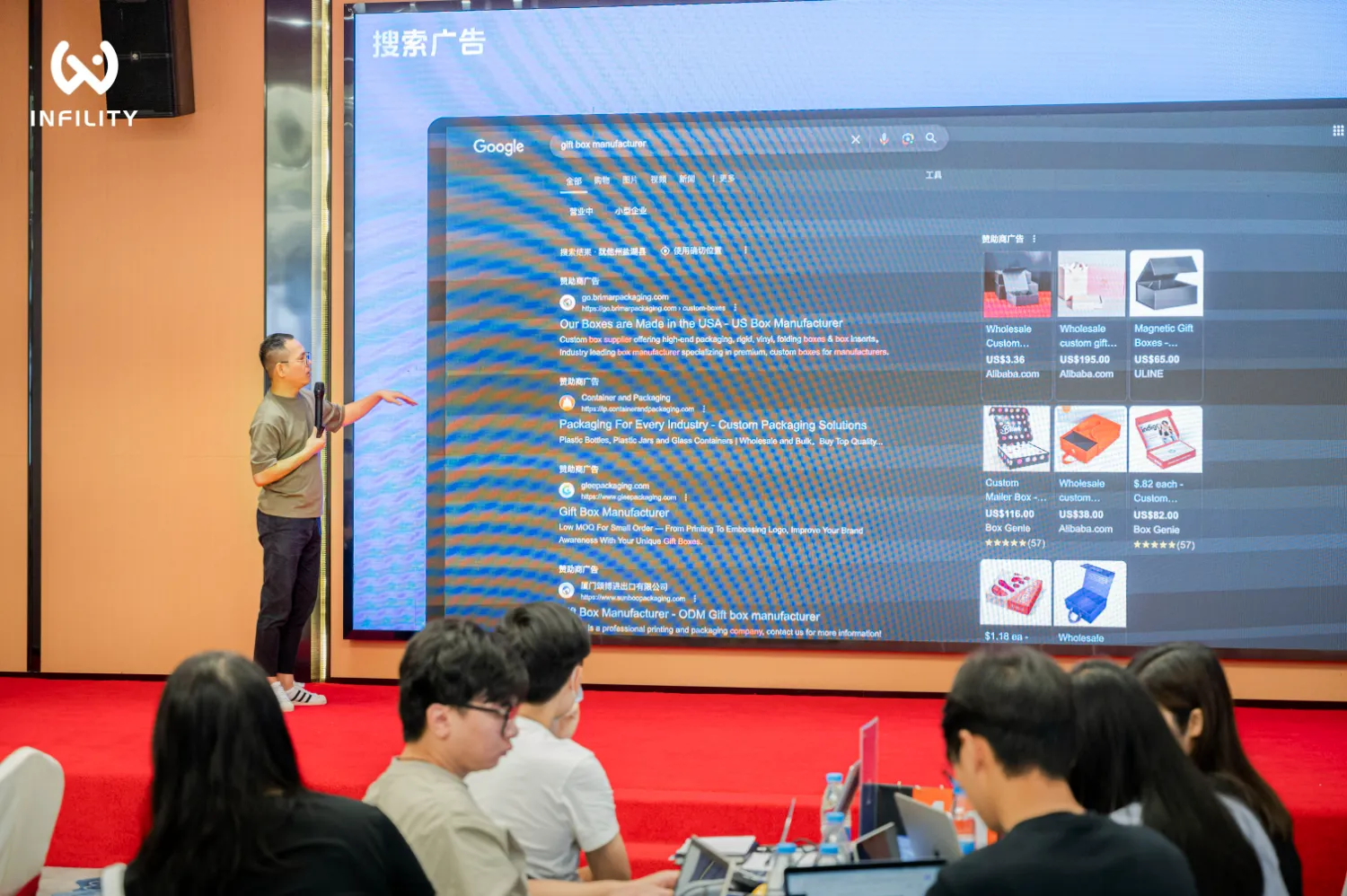 Infility 研讨会现场，讲师正在演示 Google 搜索广告效果，屏幕展示礼品盒制造商的搜索结果，台下观众专注听讲。