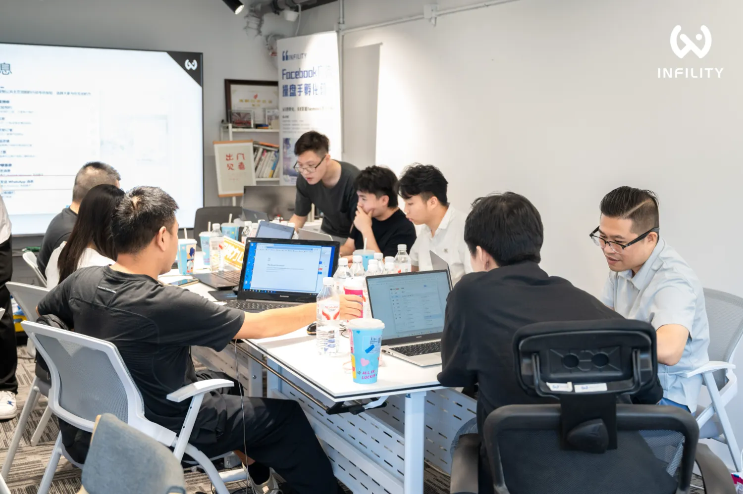 一群学员在INFINITY Facebook广告培训课上围坐使用笔记本电脑，讲师在旁指导，背景有投影屏幕。