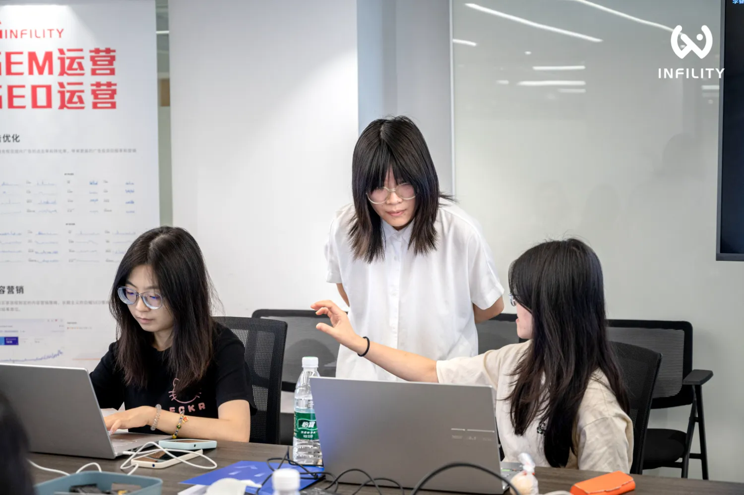 办公室内三名女性职员正围绕笔记本电脑进行业务讨论，背景可见SEM与SEO运营相关海报