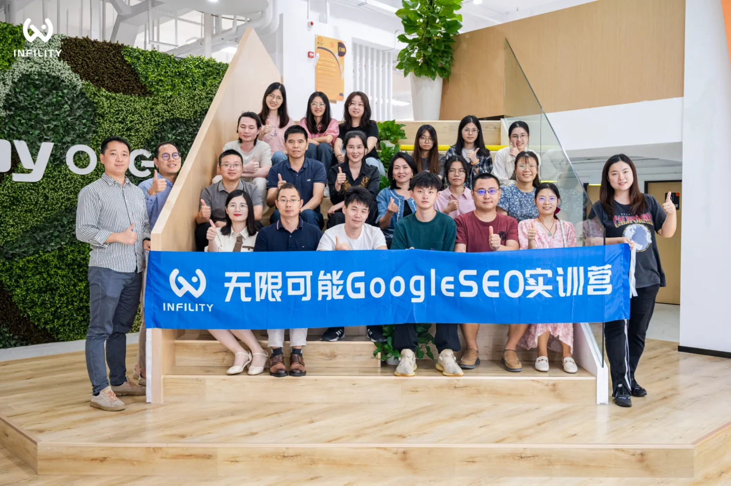 多名学员与讲师在明亮办公室的木质阶梯上合影，众人共同举着印有无限可能GoogleSEO实训营字样的蓝色横幅，微笑着竖起大拇指。