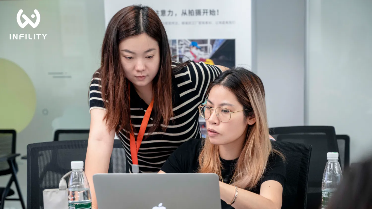 办公室内，一名穿条纹衫的女性站立俯身，与戴眼镜的坐姿女性共同专注查看笔记本电脑屏幕，背景可见INFILITY标识。