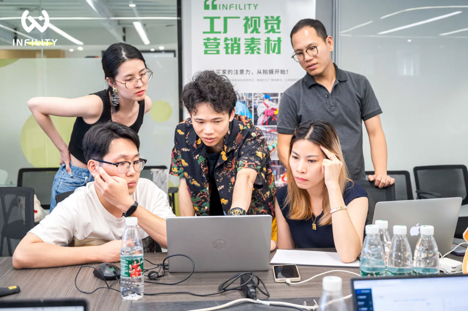 多名团队成员在办公区围坐电脑前，专注讨论与审核工厂视觉营销素材，背景墙印有相关品牌标语。