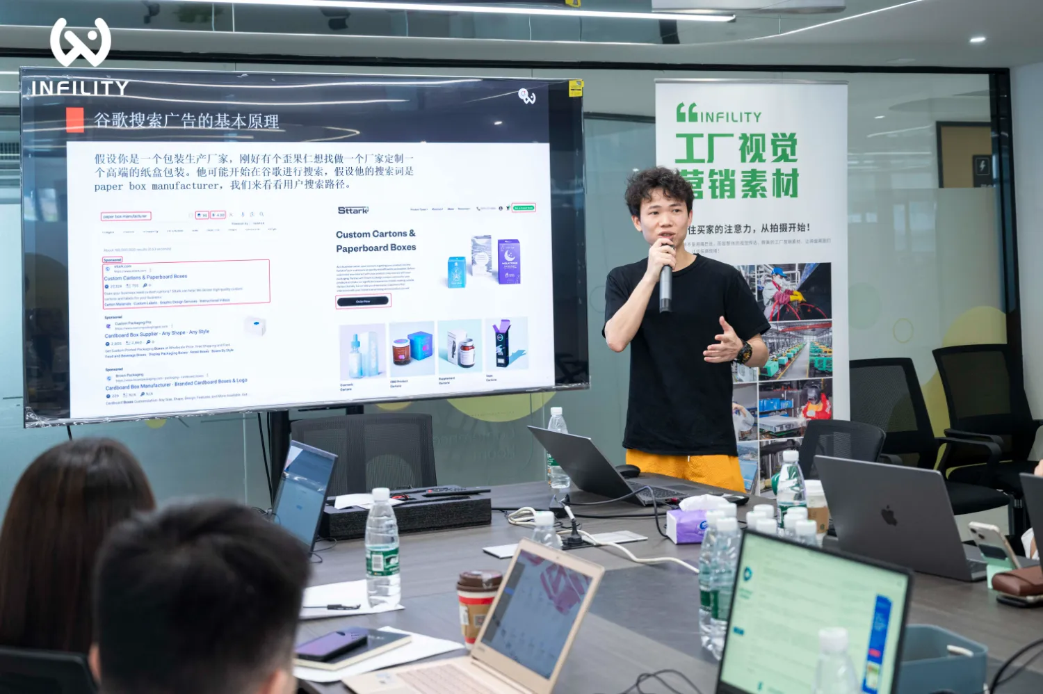 演讲者在会议室内讲解谷歌搜索广告的基本原理，屏幕展示相关案例，背景立牌显示“INFINITY工厂视觉营销素材”。