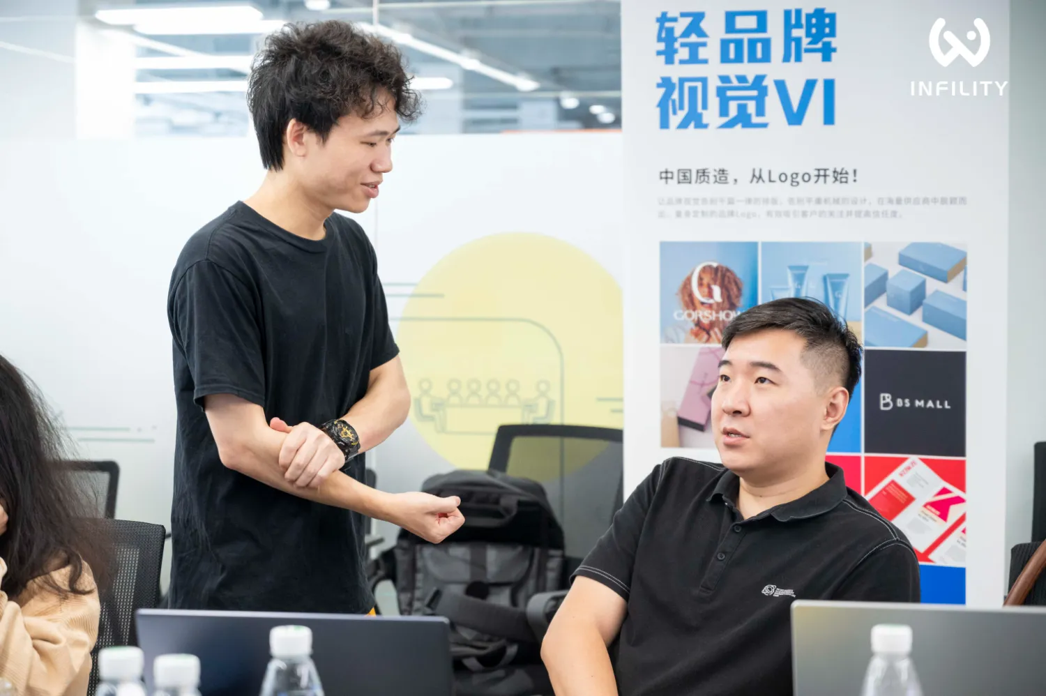 两名男士在办公室内讨论品牌视觉VI设计方案，背景为Infinity公司的宣传海报，展示了Logo设计案例。