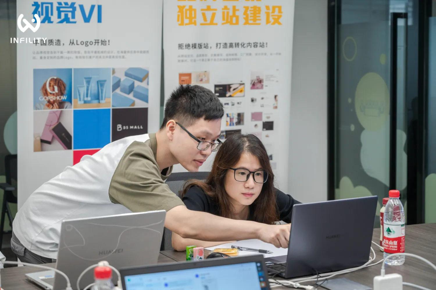 两名设计师在办公桌前共同查看笔记本电脑，背景为视觉VI与独立站建设的宣传展板。