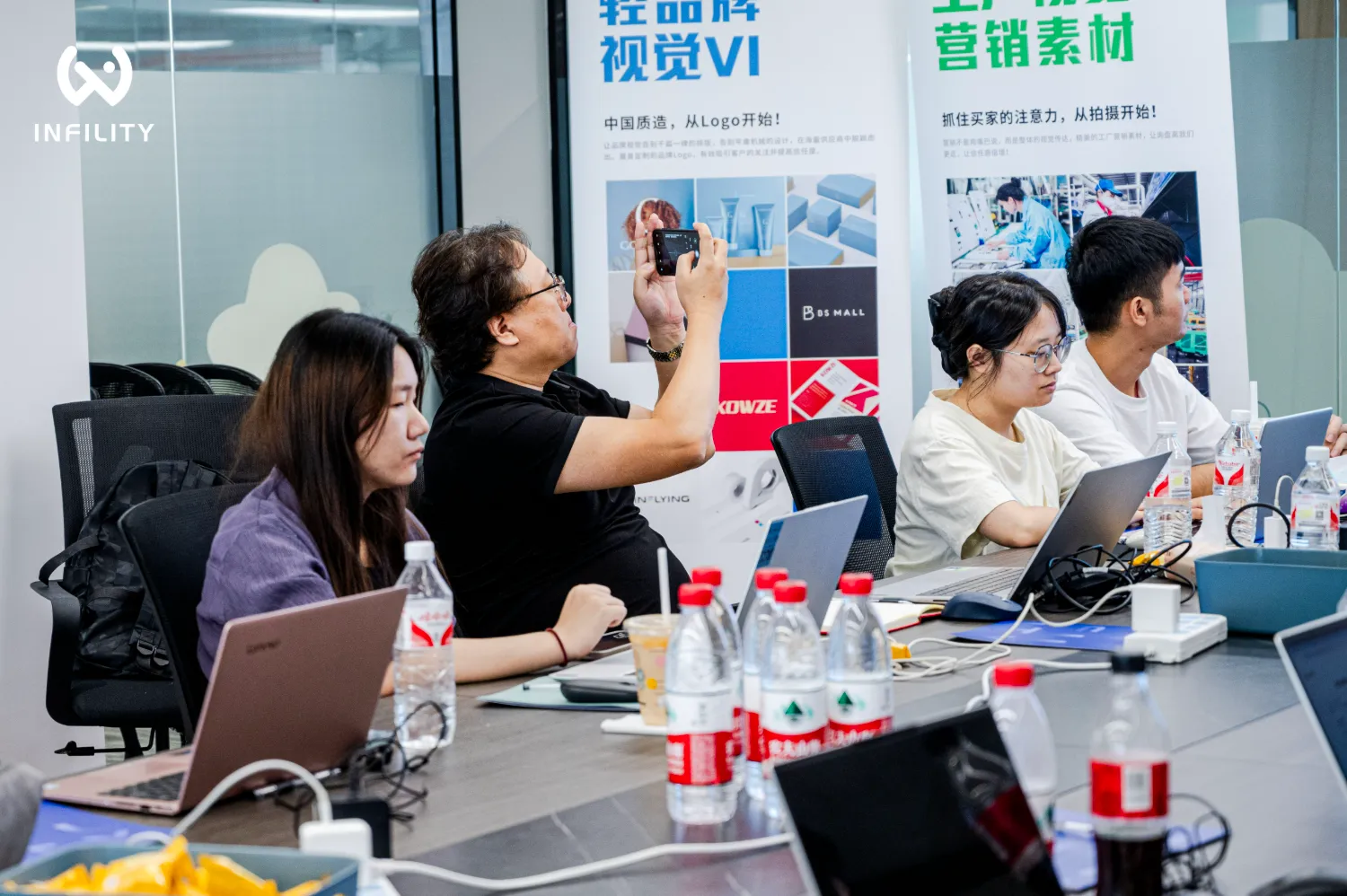 会议桌上，一名男子正举着手机拍摄背景中的品牌视觉VI与营销素材展板，其他人坐在电脑前工作。