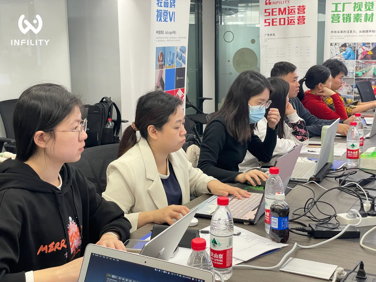 多名学员在会议室使用笔记本电脑参加培训，背景可见SEM SEO运营及工厂视觉宣传海报